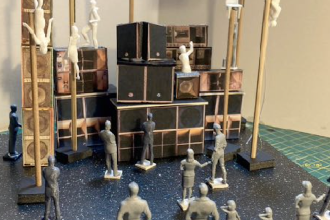 A model of the staging for Ancient Futures made from card and plastic figures of people. Bodies climb tall flag poles surrounding a high structure made of speakers while plastic figure of audience members look on.