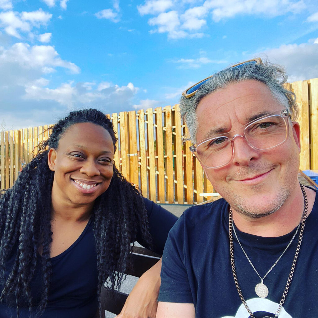 A Black woman with long black braids and a white man with curly salt and pepper hair and glasses sit next to each other, smiling broadly. A blue sky with a few clouds.