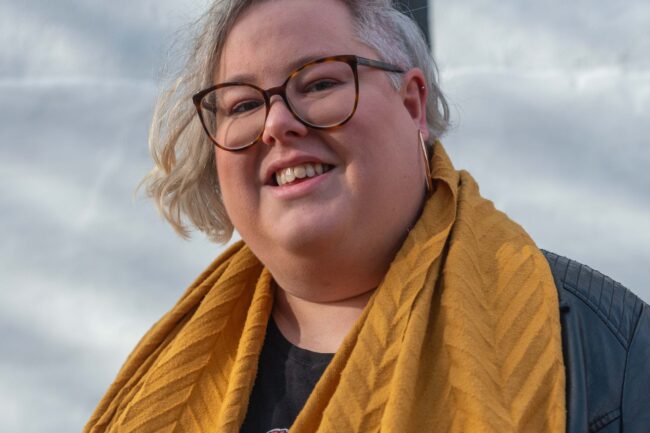 Jamie Fletcher - a white, bobbed and blonde haired trans woman with glasses, wearing black culottes and leather jacket with limited edition “Rainbow” Nike Air Max 90 trainers