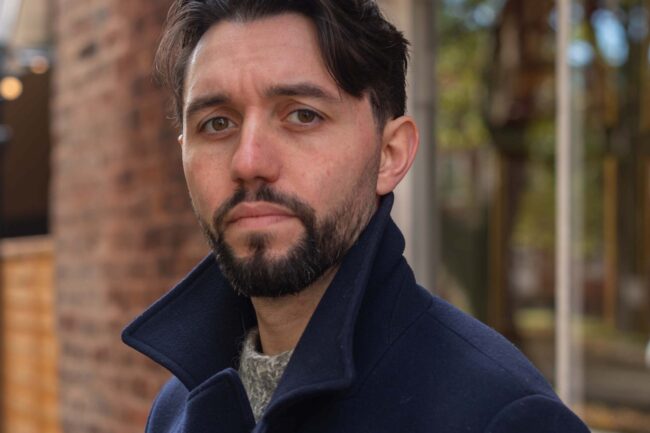Tyrrell Jones - a white, dark haired cis man with a neat beard, wearing blue jeans, blue double breasted jacket and brown desert boots