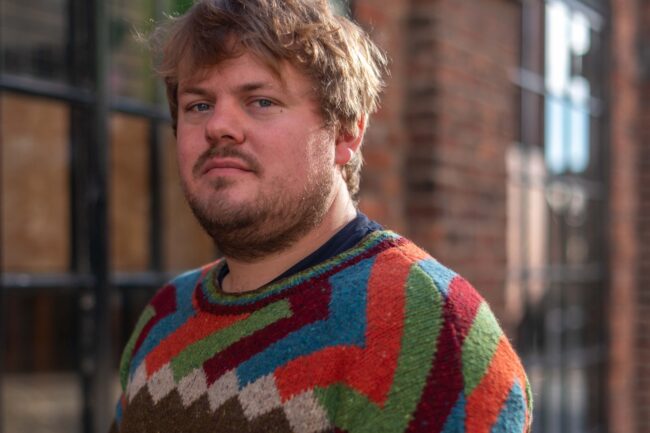 Ali Pidsley - a tousled and blonde haired cis man with a natural beard, wearing a home knitted brown jumper with orange, blue, green and maroon patterns with blue jeans and black trainers.