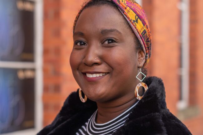 Lauren Nicole Whitter - a black cis woman with natural hair in an orange patterned headwrap, with hanging hooped earrings, wearing black faux leather dungarees over a black and white striped top, black faux leather knee length coat
