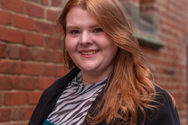 Rachael Abbey a red haired cis woman wearing green courduroy dungarees with knee length black jacket, hooped gold earrings and Adidas shell toe trainers