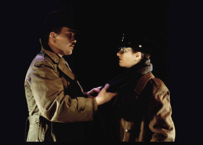 A man with a moustache holds the scarf (lovingly) of a woman in military uniform. They are actors in a play set in 1945