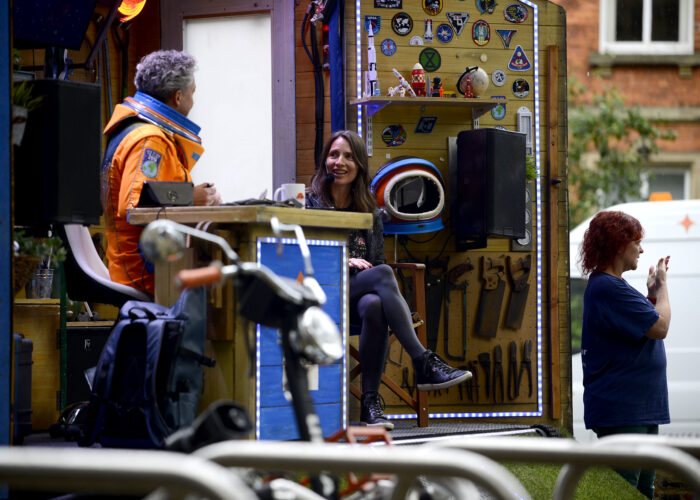 A space-suited human chats to a smiling woman in a shed. Another woman interprets their conversation into British Sign Language